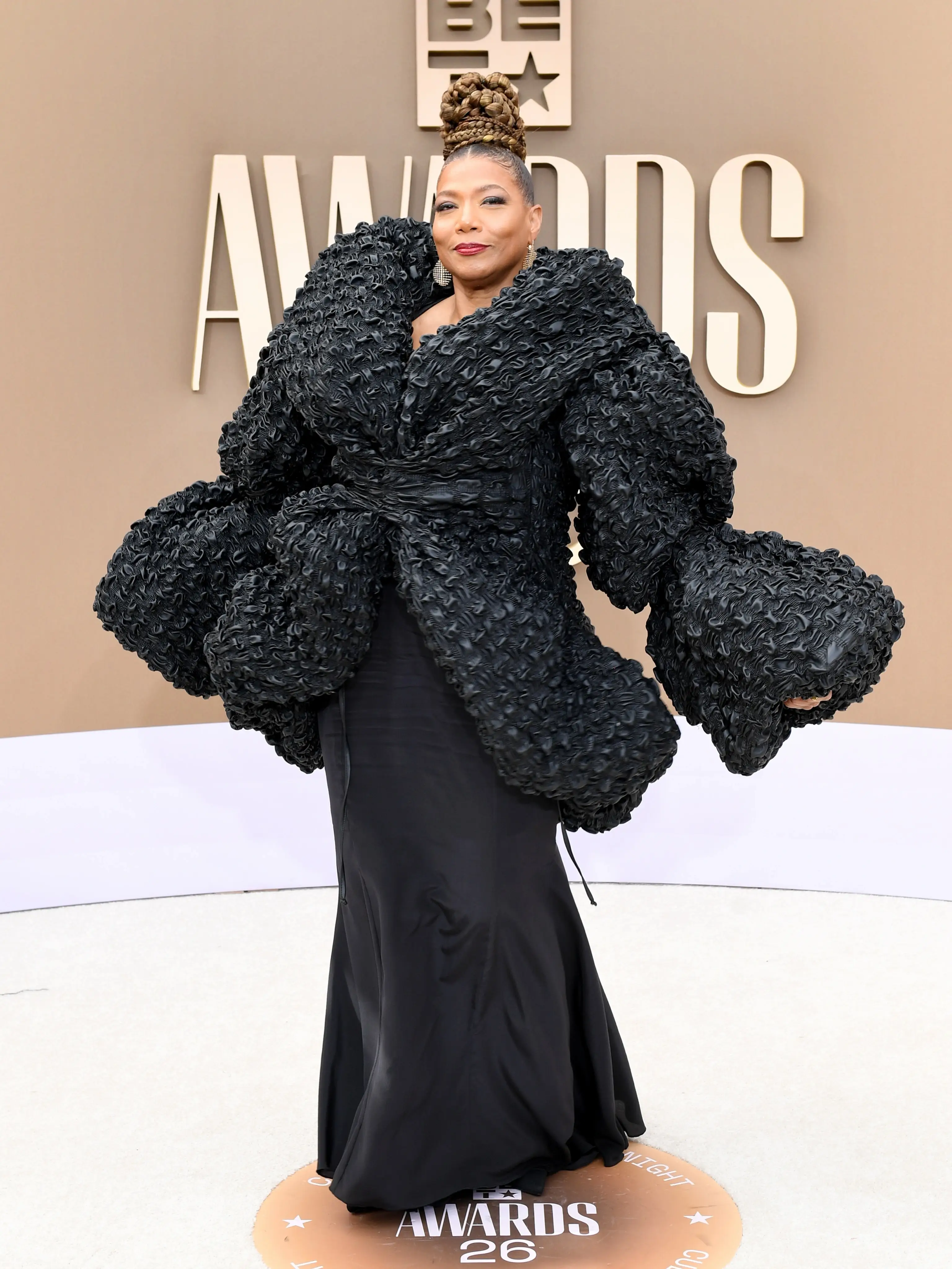 Queen Latifah poses on a red carpet wearing a dramatic, voluminous black textured wrap over a black gown at the BET Awards.