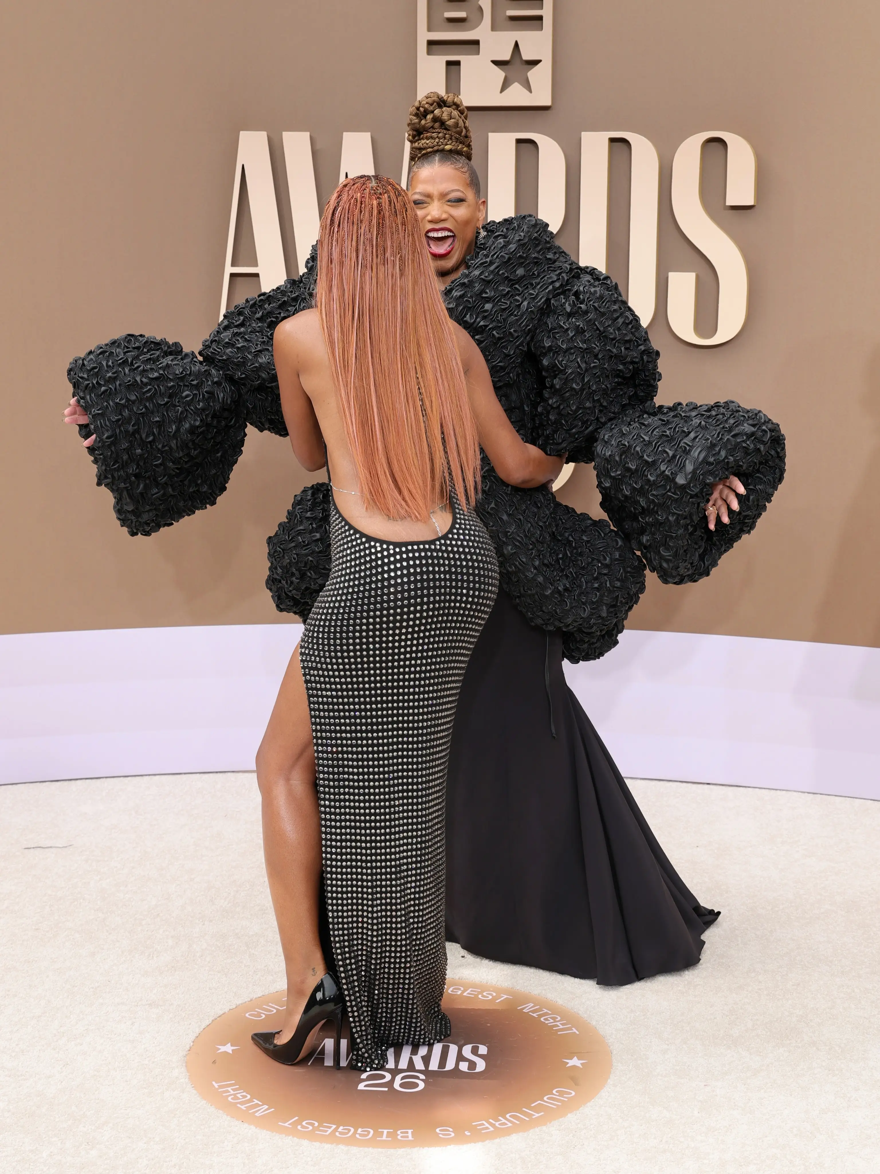 Keke Palmer, seen from behind in a studded black backless gown, greets a laughing Queen Latifah who is wearing a dramatic textured black ensemble at the BET Awards.