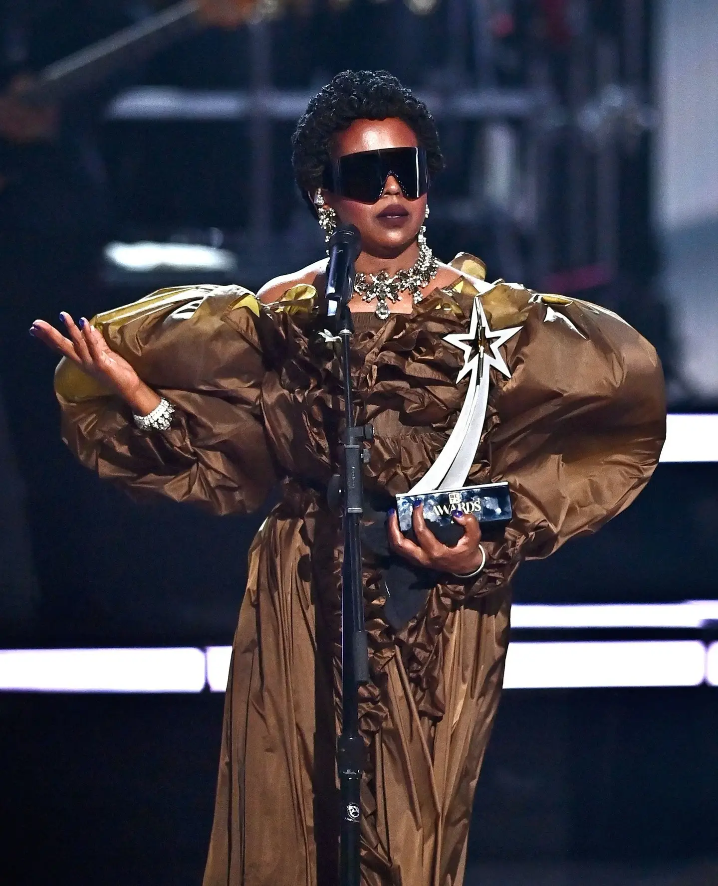 Singer Ms. Lauryn Hill stands at a microphone wearing a voluminous bronze gown, dark shield sunglasses, and heavy jewelry while holding a BET Awards trophy.