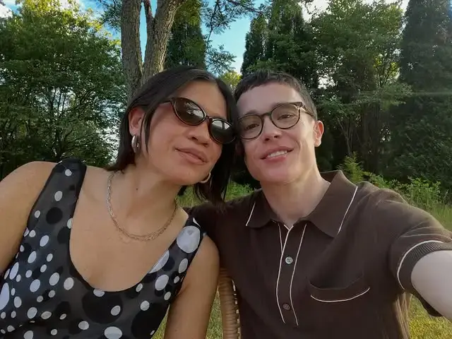 Comedian Julia Shiplett in a polka dot top and sunglasses sits close to actor Elliot Page, who wears a brown polo shirt and eyeglasses, while outdoors together.