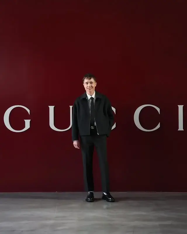 Actor Elliot Page smiles while posing in a black jacket, tie, and dress pants in front of a large red Gucci backdrop.