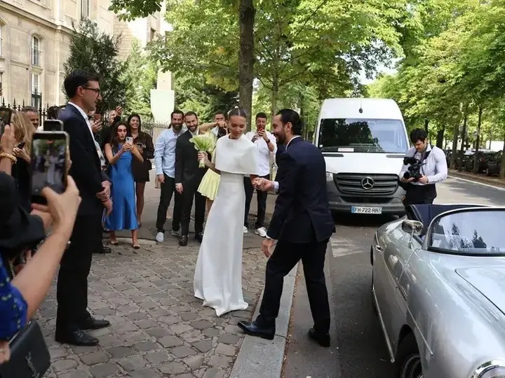 Model Thylane Blondeau in a white wedding gown holds hands with her husband Ben Attal as they walk toward a vintage convertible car.
