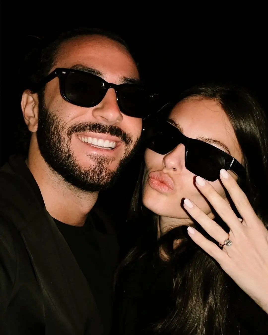 Model Thylane Blondeau makes a kissy face and flashes her engagement ring alongside her smiling husband Ben Attal while both wear black sunglasses.