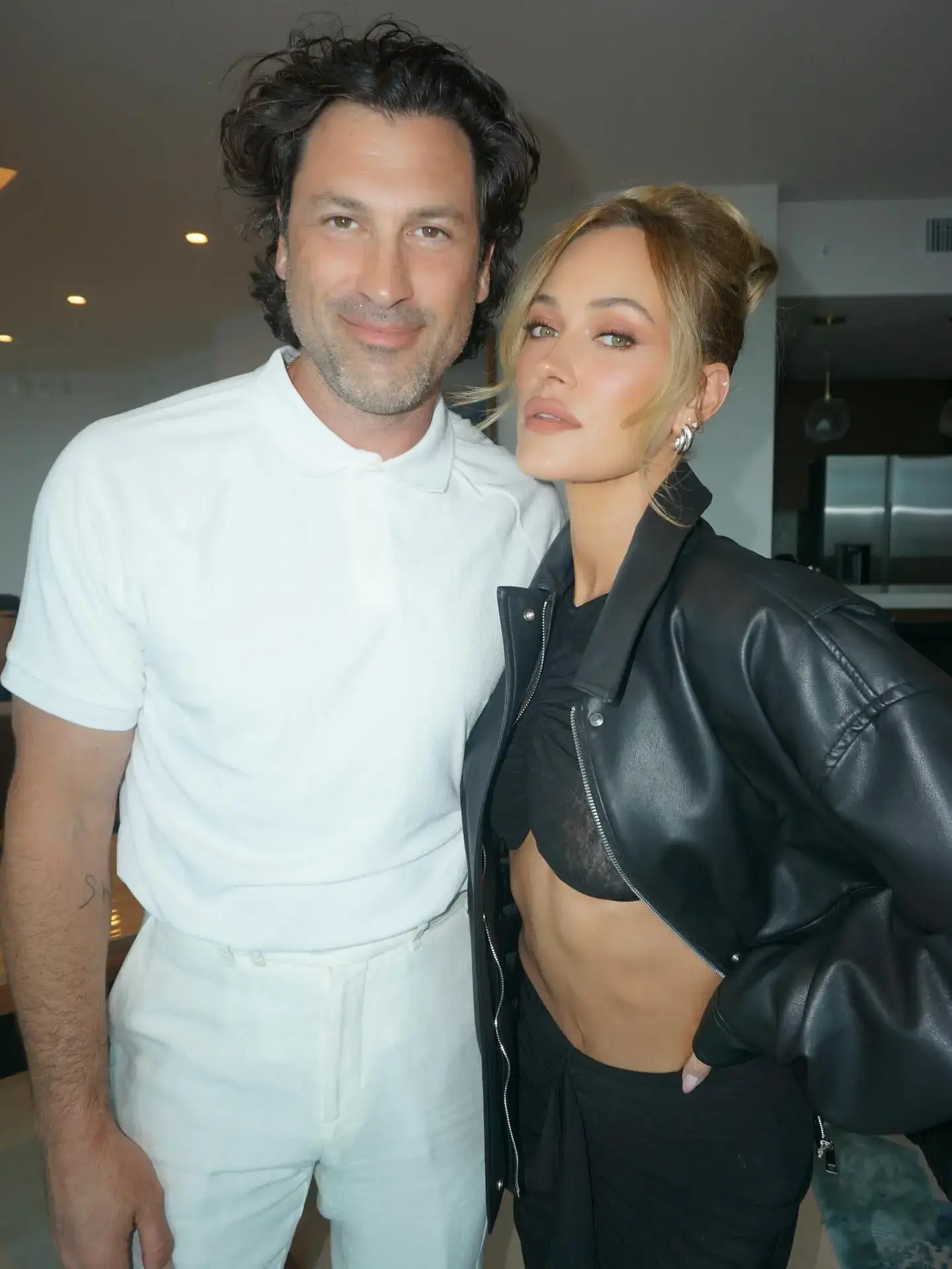 Maks Chmerkovskiy in an all-white polo outfit stands next to Peta Murgatroyd who is wearing a black leather jacket and crop top indoors.