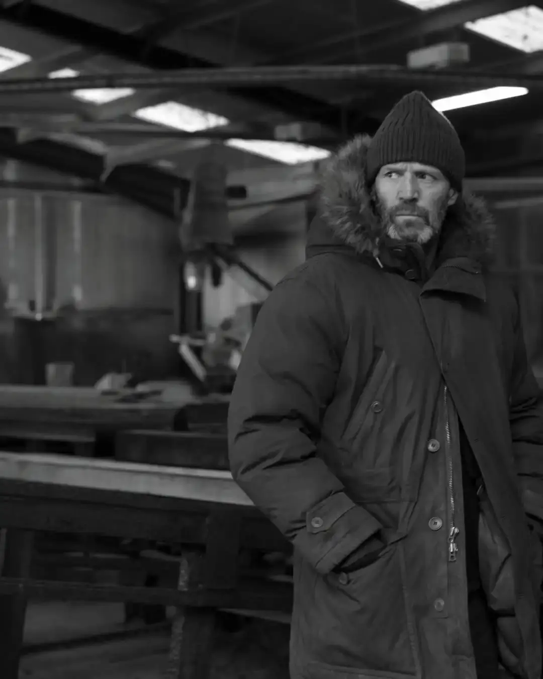 A black and white image of a bearded Jason Statham wearing a fur-lined winter parka and beanie while looking to the side inside a workshop.