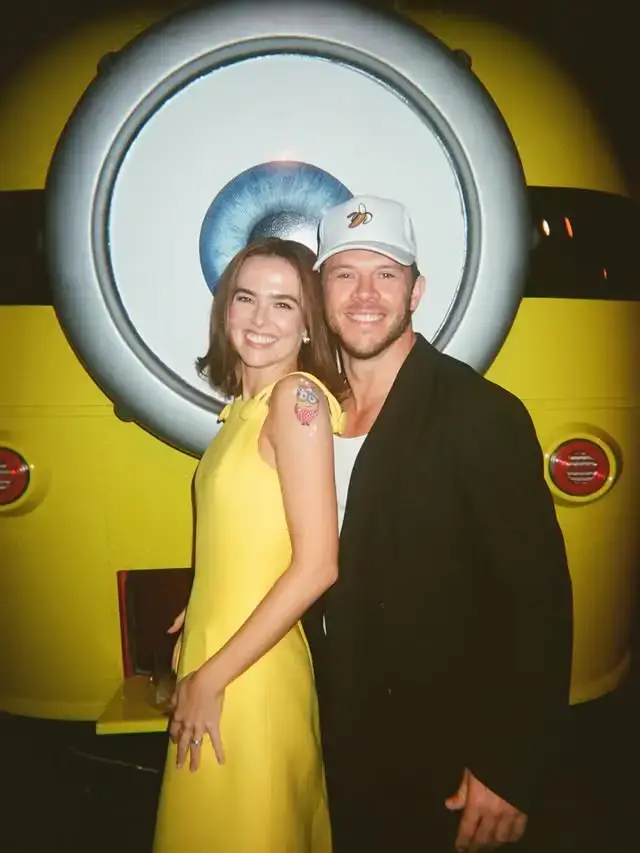 Actress Zoey Deutch in a bright yellow dress stands alongside her boyfriend Jimmy Tatro, who wears a black jacket and white banana hat, in front of a giant Minion backdrop.