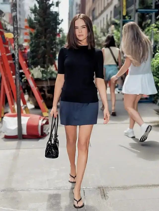 Actress Zoey Deutch walks down a city sidewalk wearing a short-sleeved black top, blue mini skirt, and sandals while holding a handbag.