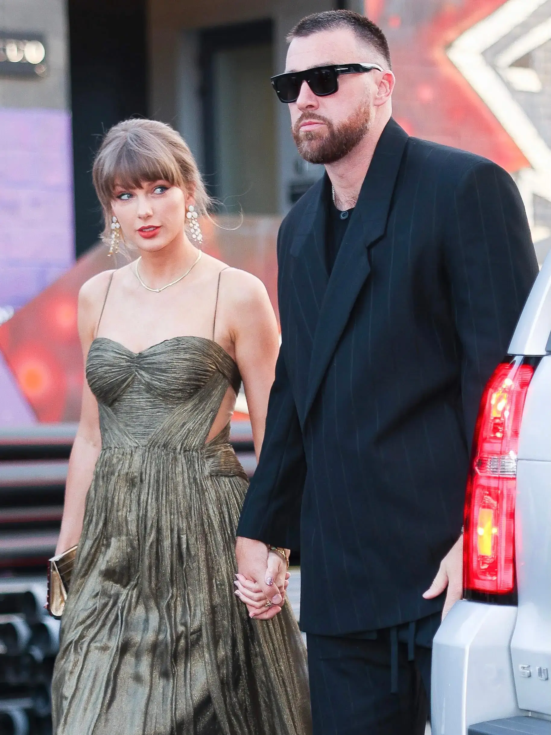 Taylor Swift in a metallic gold gown holds hands with Travis Kelce, who wears a black pinstripe suit and sunglasses, as they walk outdoors.