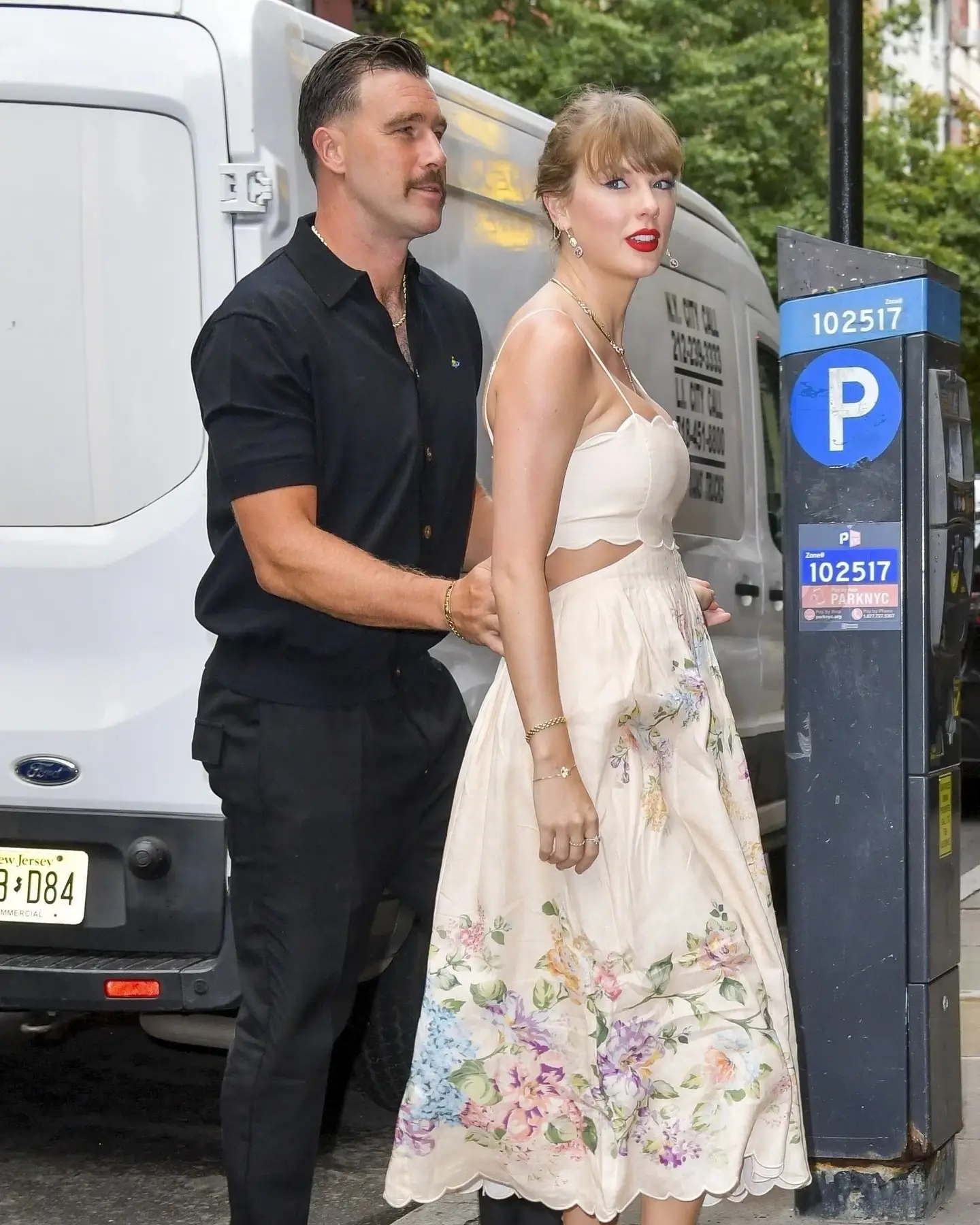 Taylor Swift in a cream-colored floral dress walks past a parking meter while Travis Kelce, in a black shirt and trousers, walks close behind her.