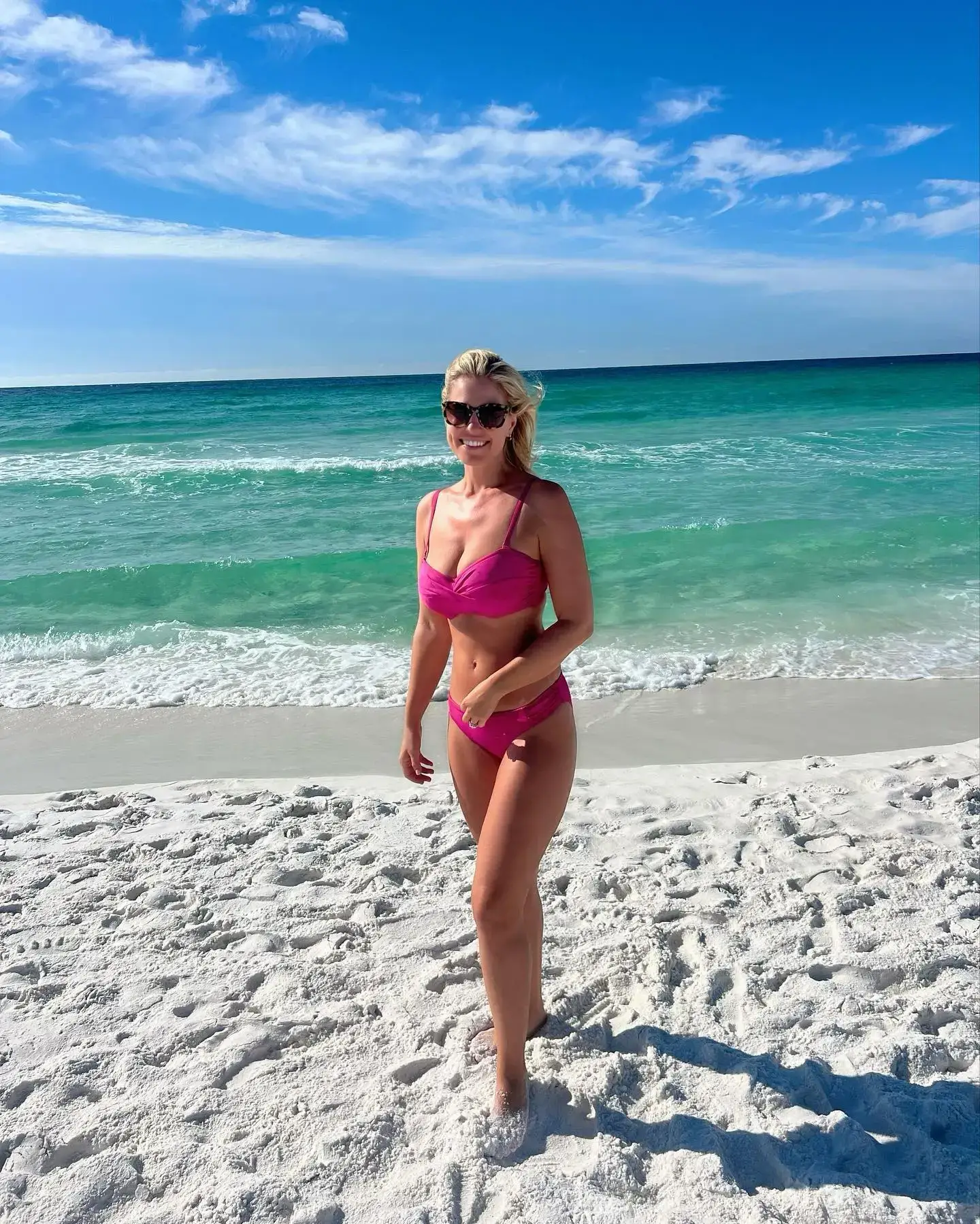 Singer-songwriter Jillian Cardarelli smiles while posing in a pink bikini and sunglasses on a white sand beach with turquoise ocean water behind her.