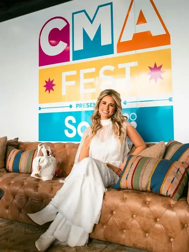 Singer-songwriter Jillian Cardarelli smiles while sitting on a brown leather tufted couch in a white lace dress with a colorful CMA Fest backdrop behind her.