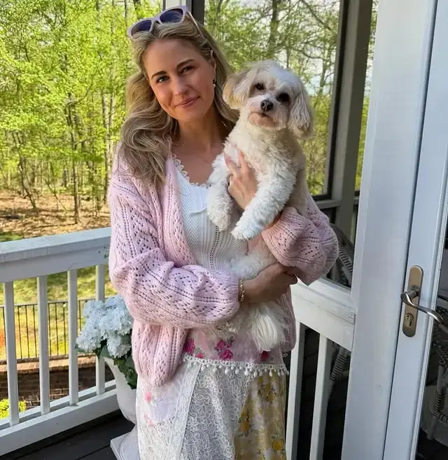 Singer-songwriter Jillian Cardarelli wears a pink knit cardigan and holds a small, fluffy white dog while standing on a porch.