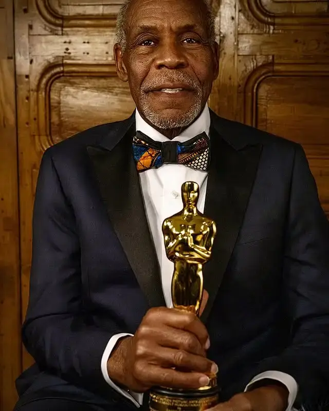 Actor Danny Glover wears a navy blue tuxedo and a patterned bow tie while holding an Oscar statuette in front of a wooden wall.