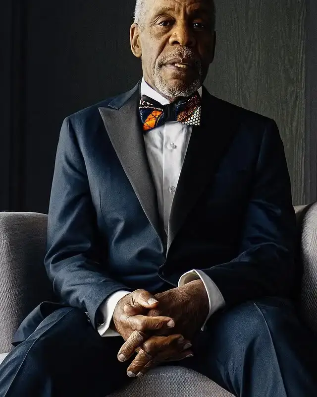 Actor Danny Glover sits in an armchair wearing a navy blue tuxedo and a patterned bow tie with his hands clasped.