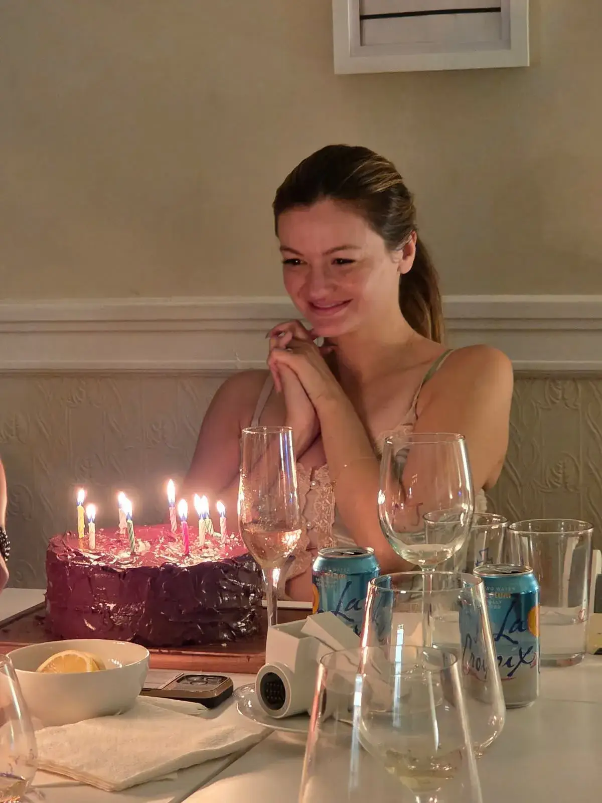 Leah McKendrick smiles with her hands clasped together behind a chocolate birthday cake topped with lit candles at a dinner table.