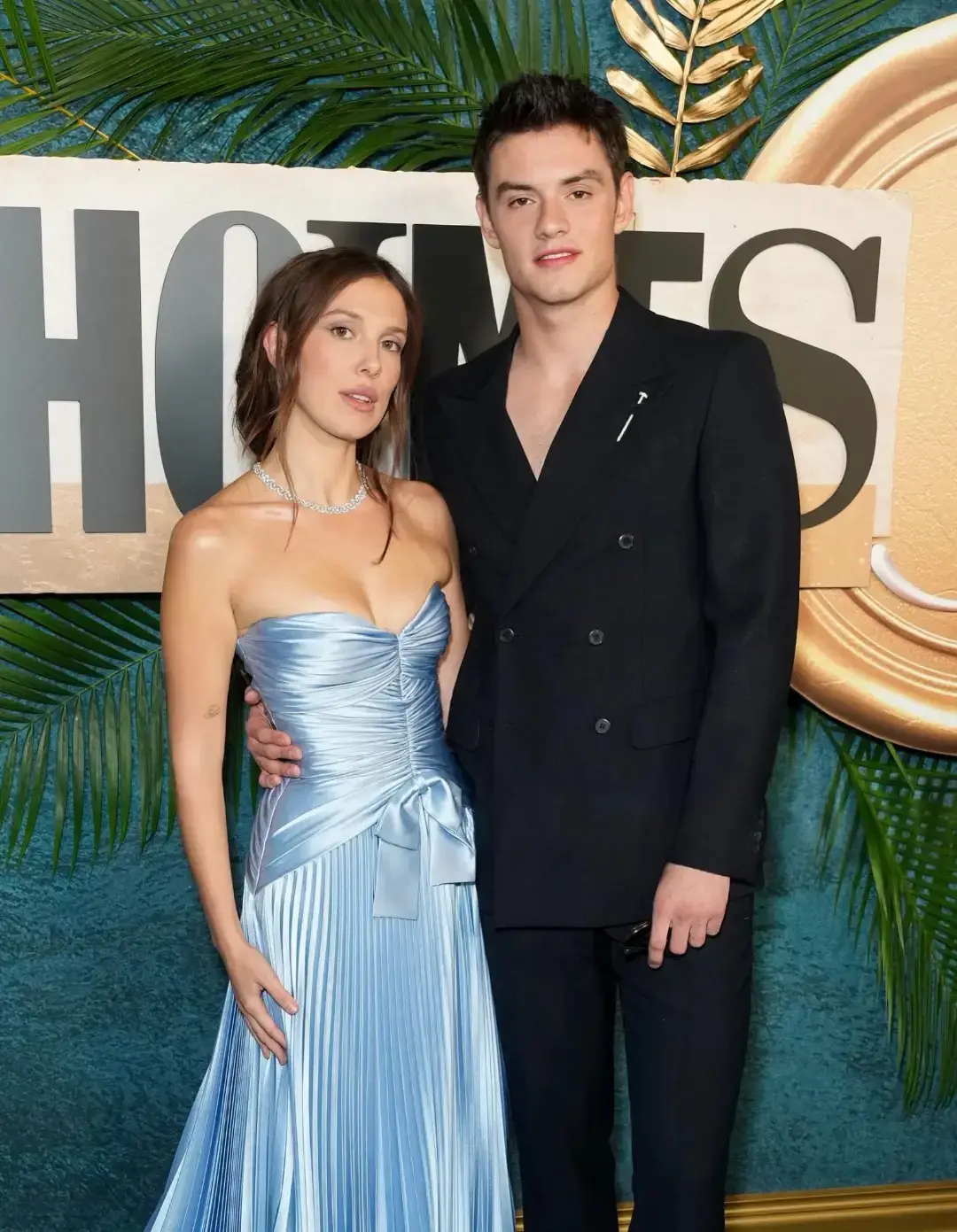 Millie Bobby Brown in a strapless light blue pleated gown poses alongside Louis Partridge in a black double-breasted suit in front of an Enola Holmes backdrop.
