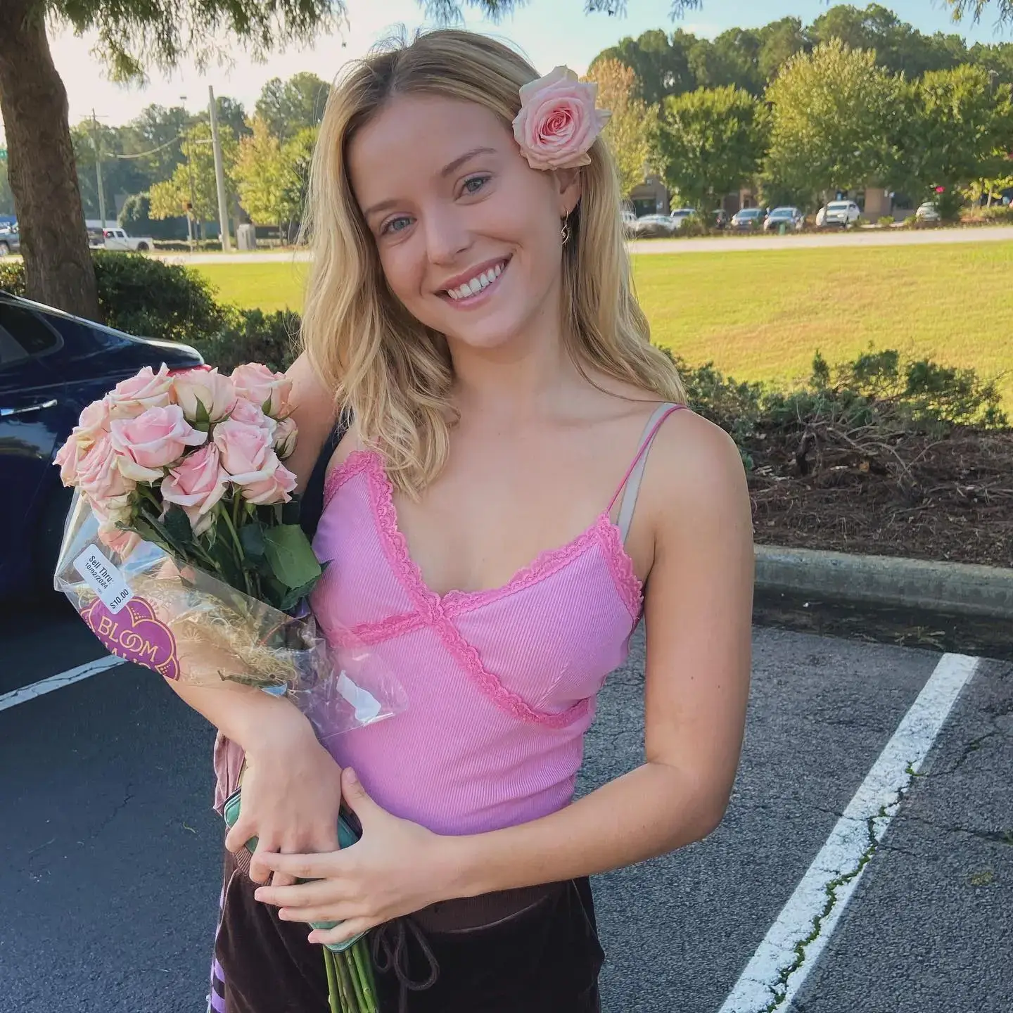 A smiling blonde woman identified as Lexi Minetree wears a pink lace-trimmed tank top and stands in a parking lot holding a bouquet of pink roses with one tucked behind her ear.