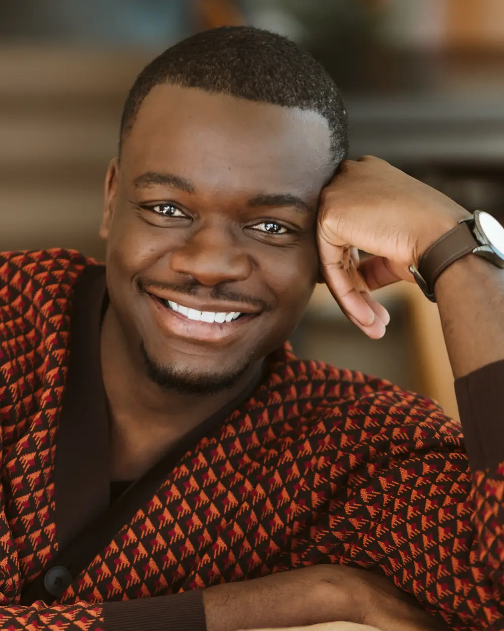 A close-up headshot of Tapiwa Soropa smiling warmly with his head resting on his hand while wearing a patterned brown and orange cardigan.