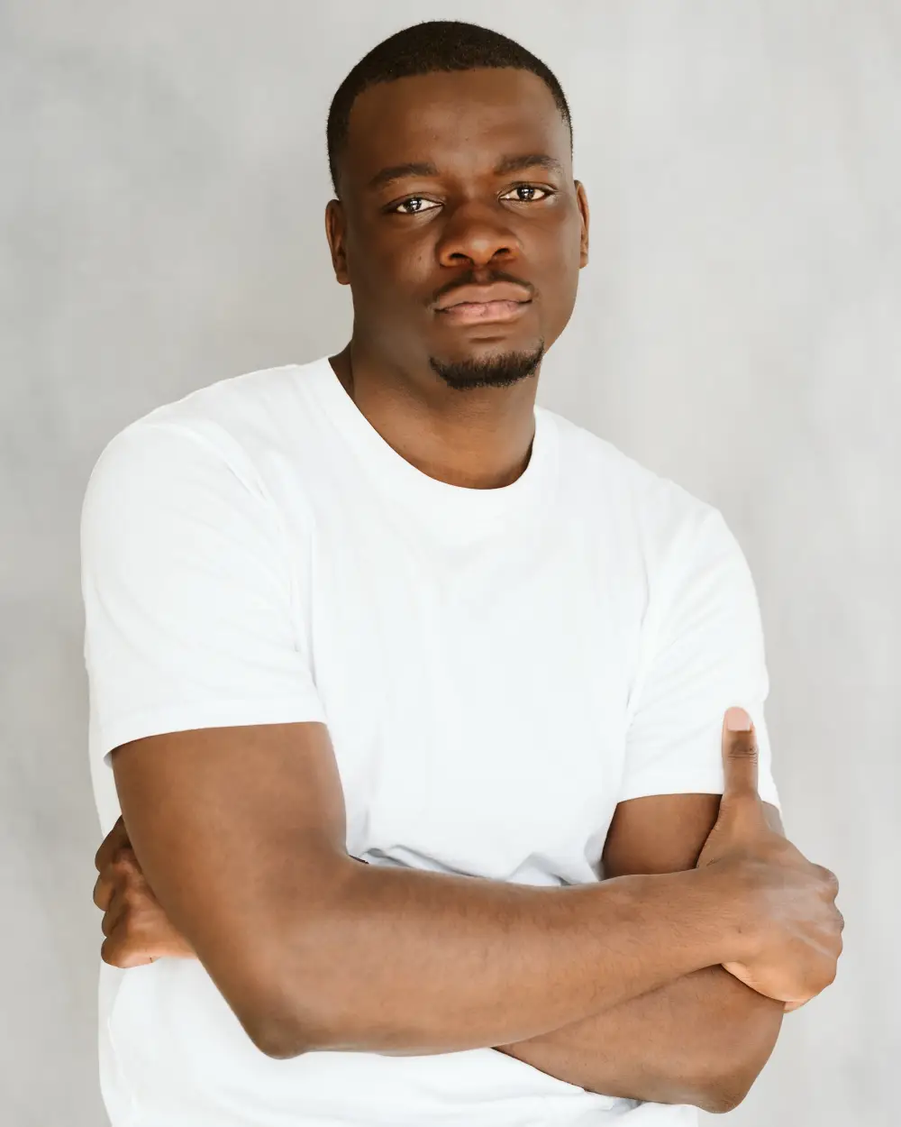 A waist-up shot of Tapiwa Soropa looking forward with a serious expression and arms crossed while wearing a white t-shirt against a plain grey background.