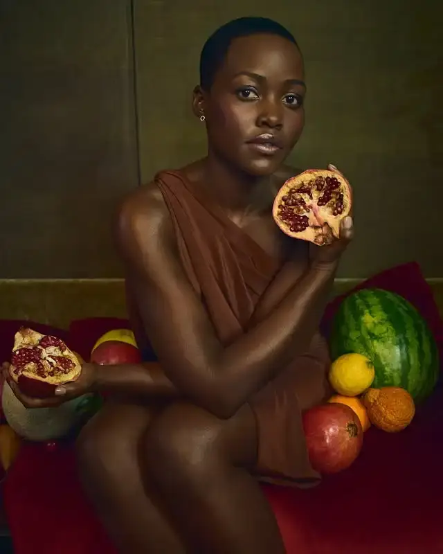 Actress Lupita Nyong’o sits wearing a brown dress while holding a split pomegranate in each hand surrounded by whole fruits