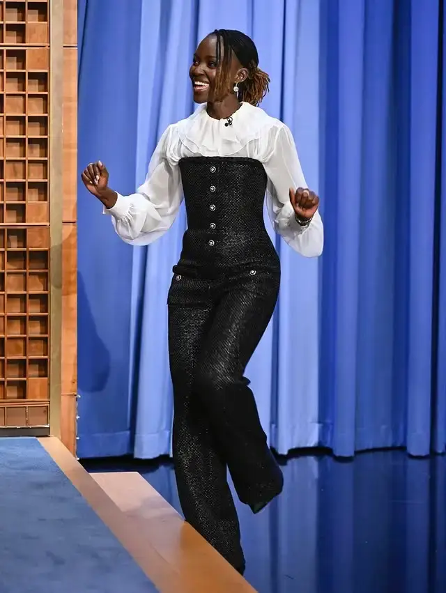 Actress Lupita Nyong’o smiles and dances onto a late-night talk show stage wearing a high-waisted black textured jumpsuit over a ruffled white blouse.