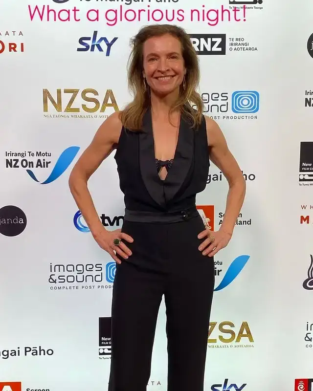 Actress Tandi Wright smiles with her hands on her hips while wearing a black sleeveless jumpsuit on a red carpet with sponsor logos.