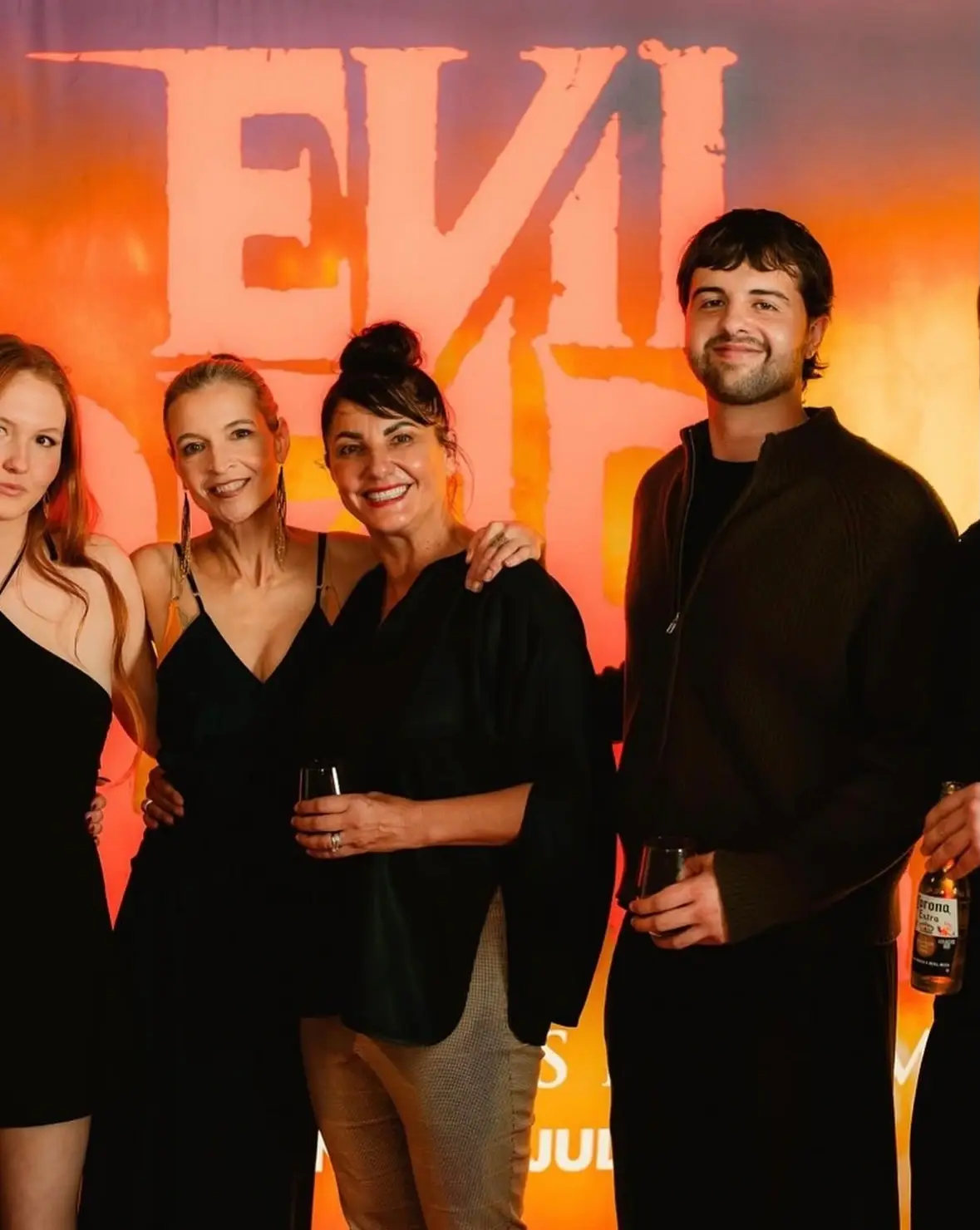 Actress Tandi Wright poses with director Lee Cronin, actress Lily Sullivan, and a guest in front of a glowing orange "Evil Dead Rise" backdrop.