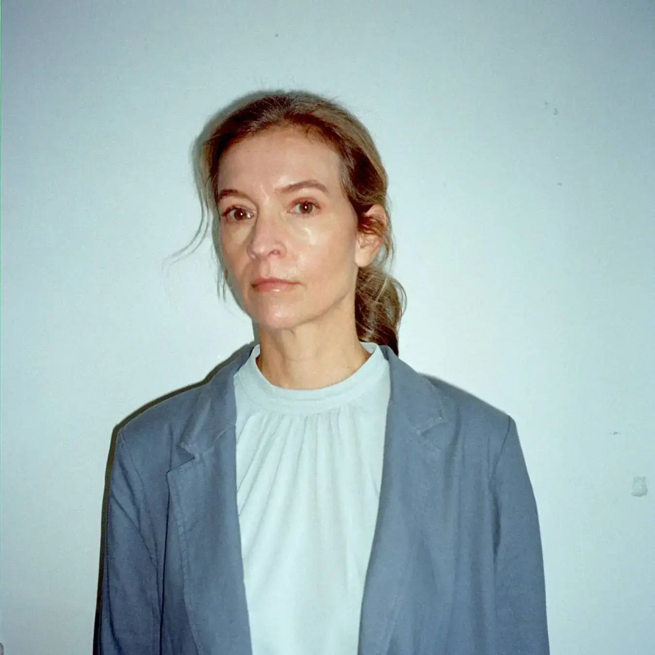 A portrait of actress Tandi Wright with a neutral expression, wearing a light blue top and a dusty blue blazer against a plain light wall.