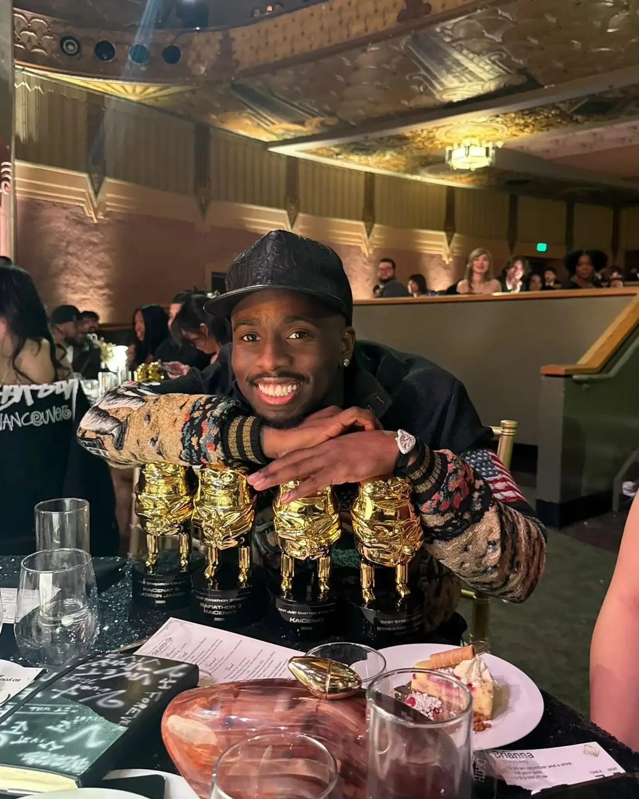 Kai Cenat smiles broadly while leaning on four golden trophies at a banquet table during an awards ceremony.