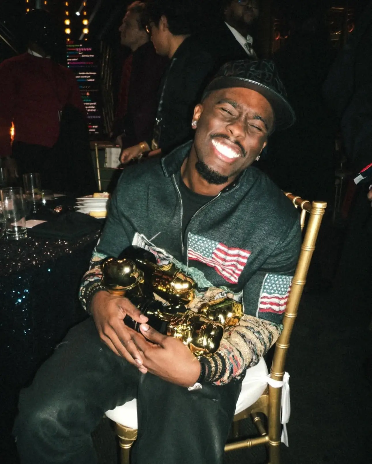 Kai Cenat laughs with joy while cradling an armful of golden trophies in his lap at an awards event banquet.