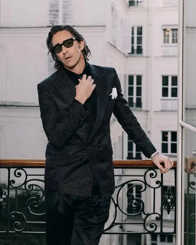 Actor Lee Pace poses on a balcony overlooking buildings while wearing a patterned black blazer, a black shirt, and dark sunglasses.