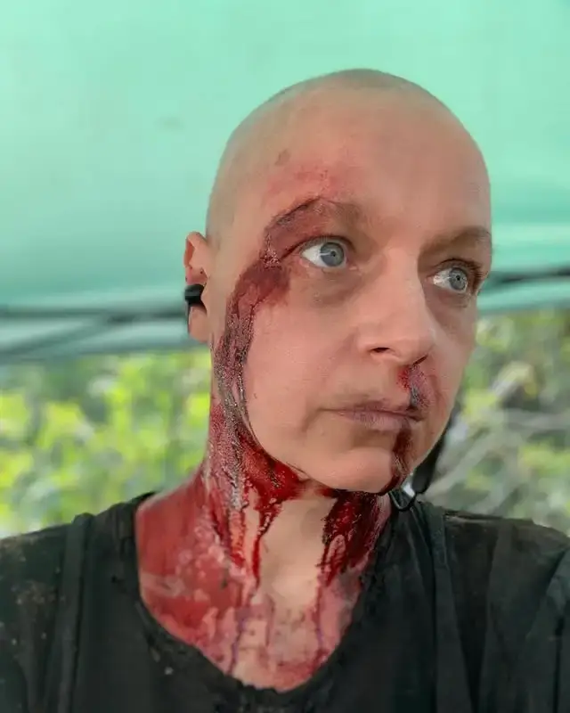 Close-up selfie of a bald Samantha Morton looking off-camera with realistic special effects makeup simulating severe bleeding from her forehead, eye, and neck.