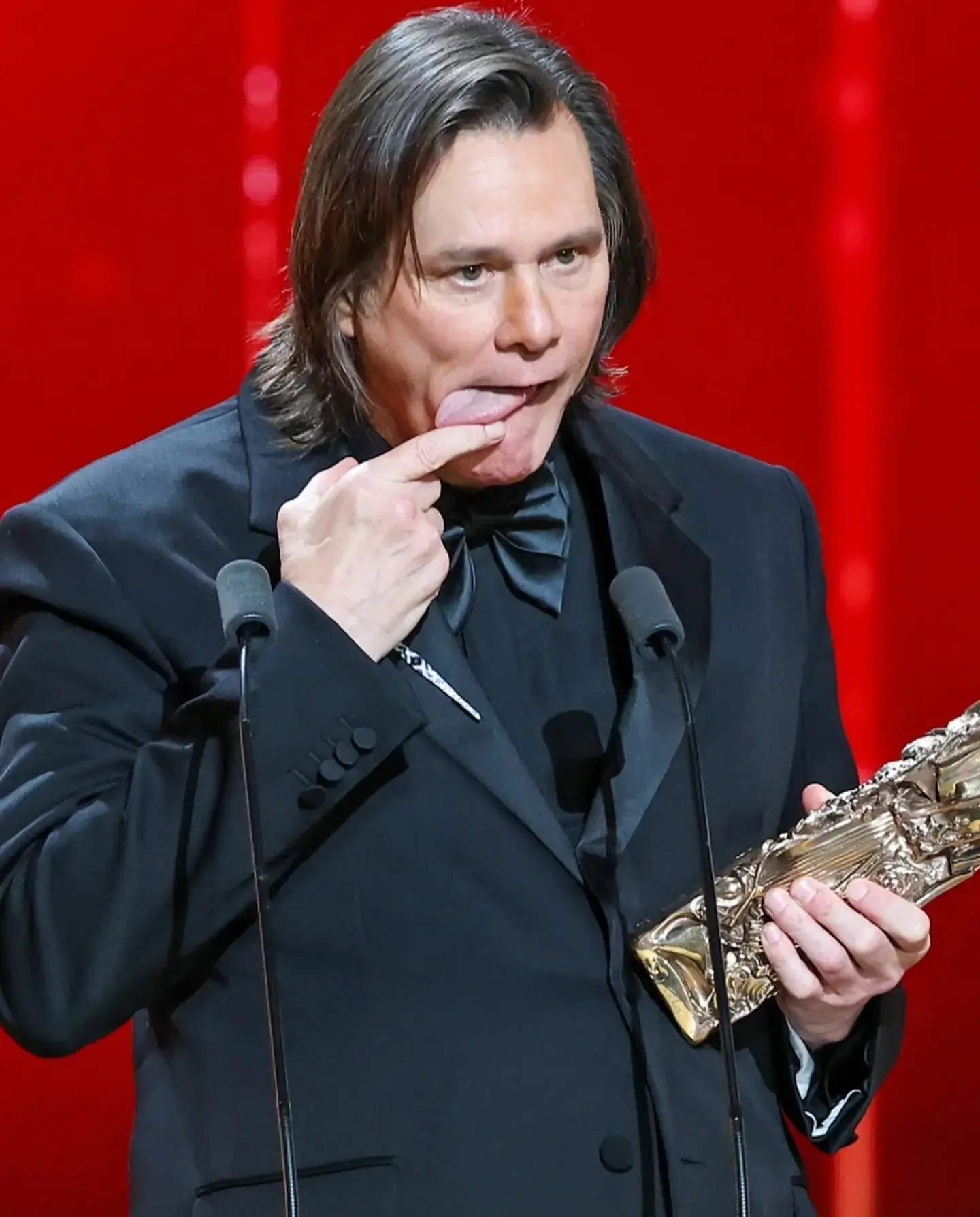 A close-up shot of Jim Carrey making a funny face by touching his tongue with his finger while holding his golden trophy at the 51st Cesar Film Awards.