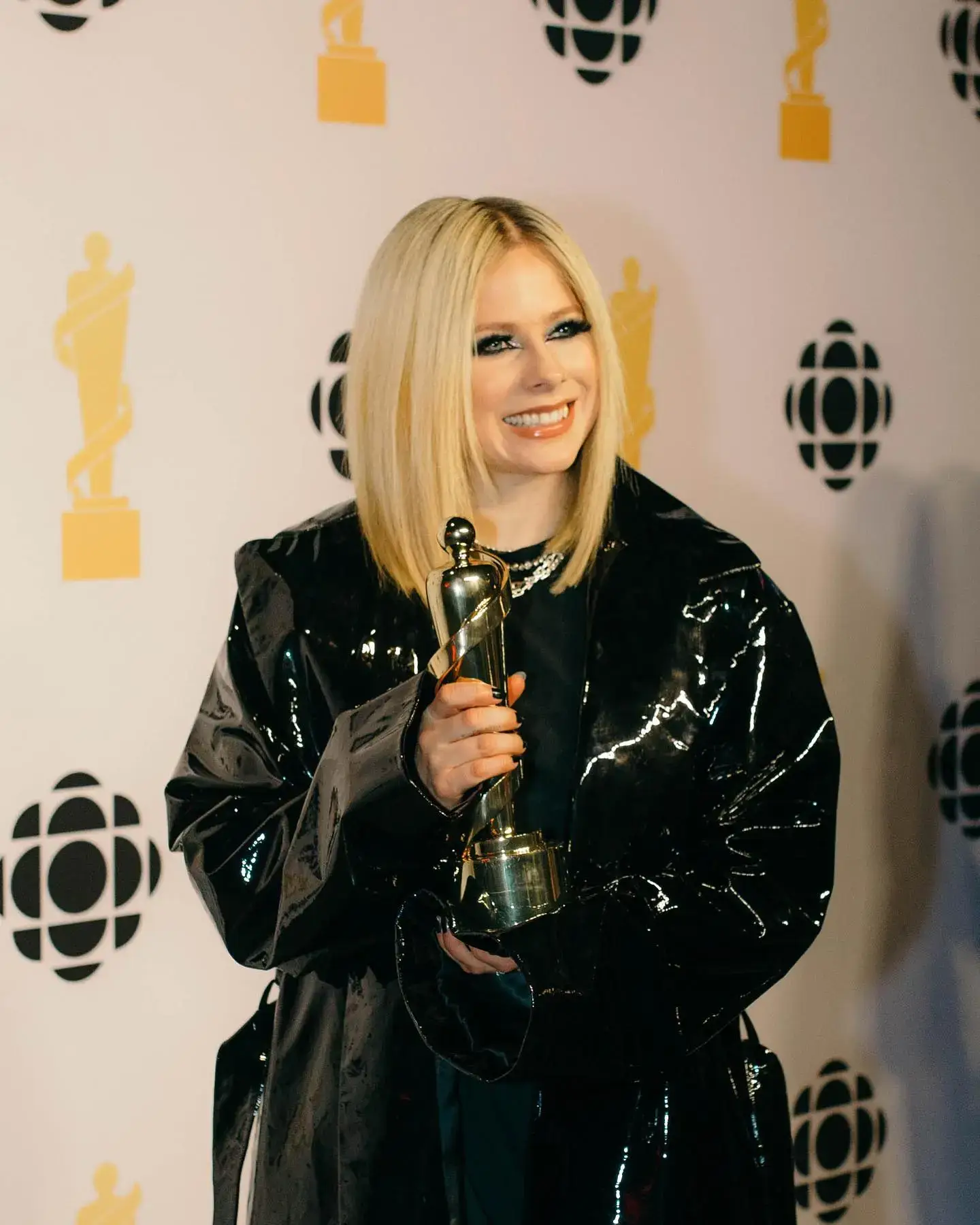 Avril Lavigne smiles while holding a golden trophy in a shiny black coat against a press backdrop with CBC logos.