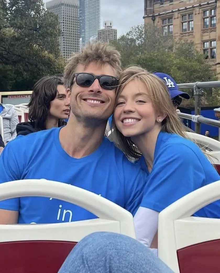 Glen Powell and Sydney Sweeney lean close and smile brightly while wearing matching blue T-shirts on an open-top tour bus.