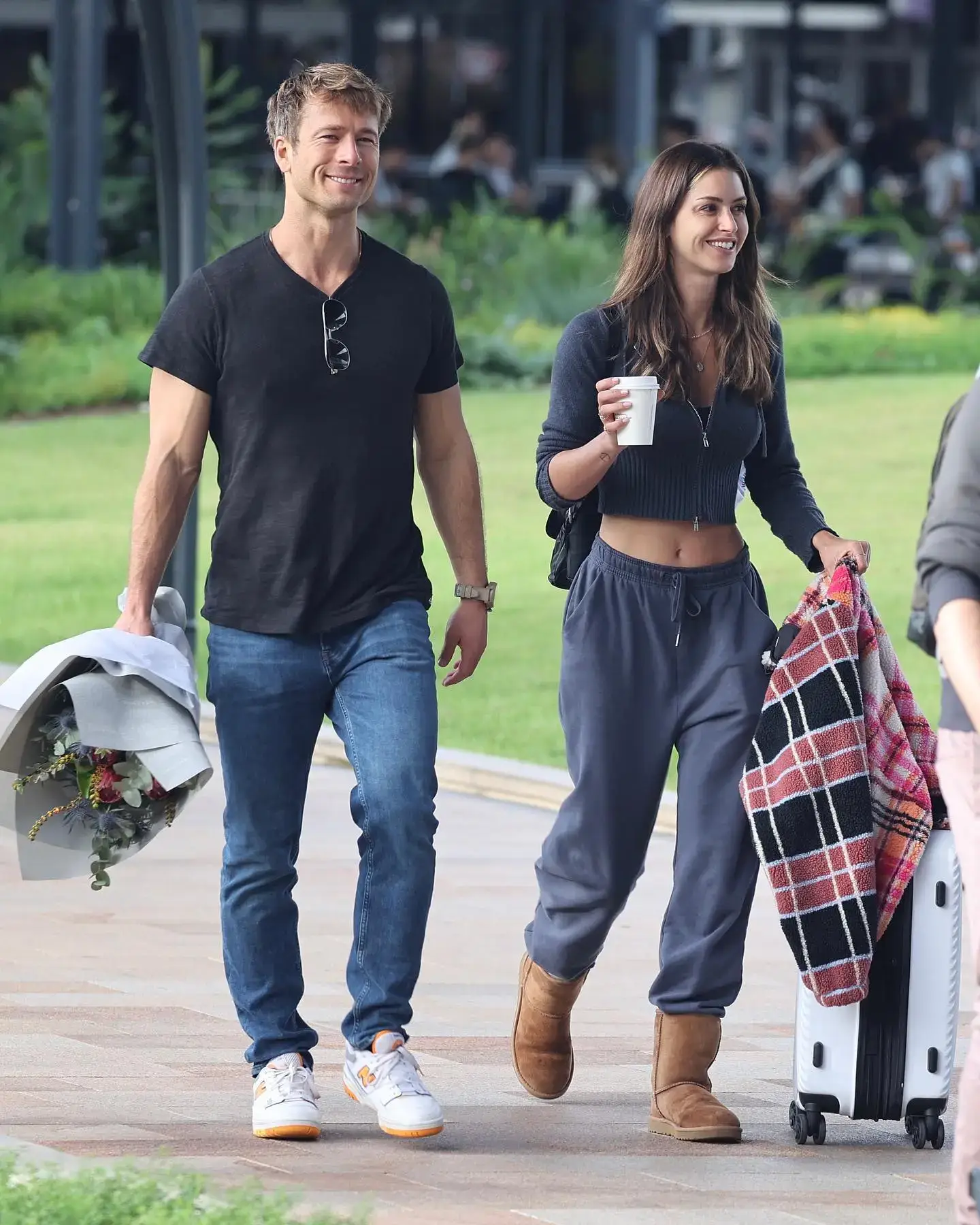 Glen Powell walks and smiles next to Gigi Paris, who holds a coffee cup and pulls a suitcase with a plaid blanket on it.