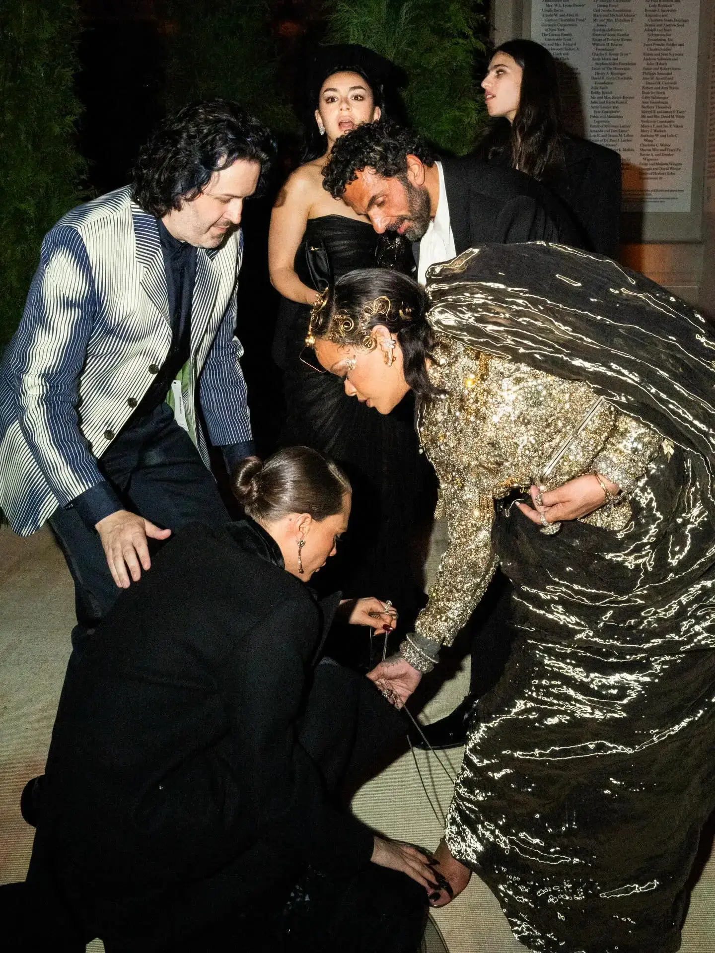 Rihanna stands in a sculptural gold Maison Margiela gown while her design team adjusts the hem, with Charli XCX reacting in surprise in the background at the 2026 Met Gala.