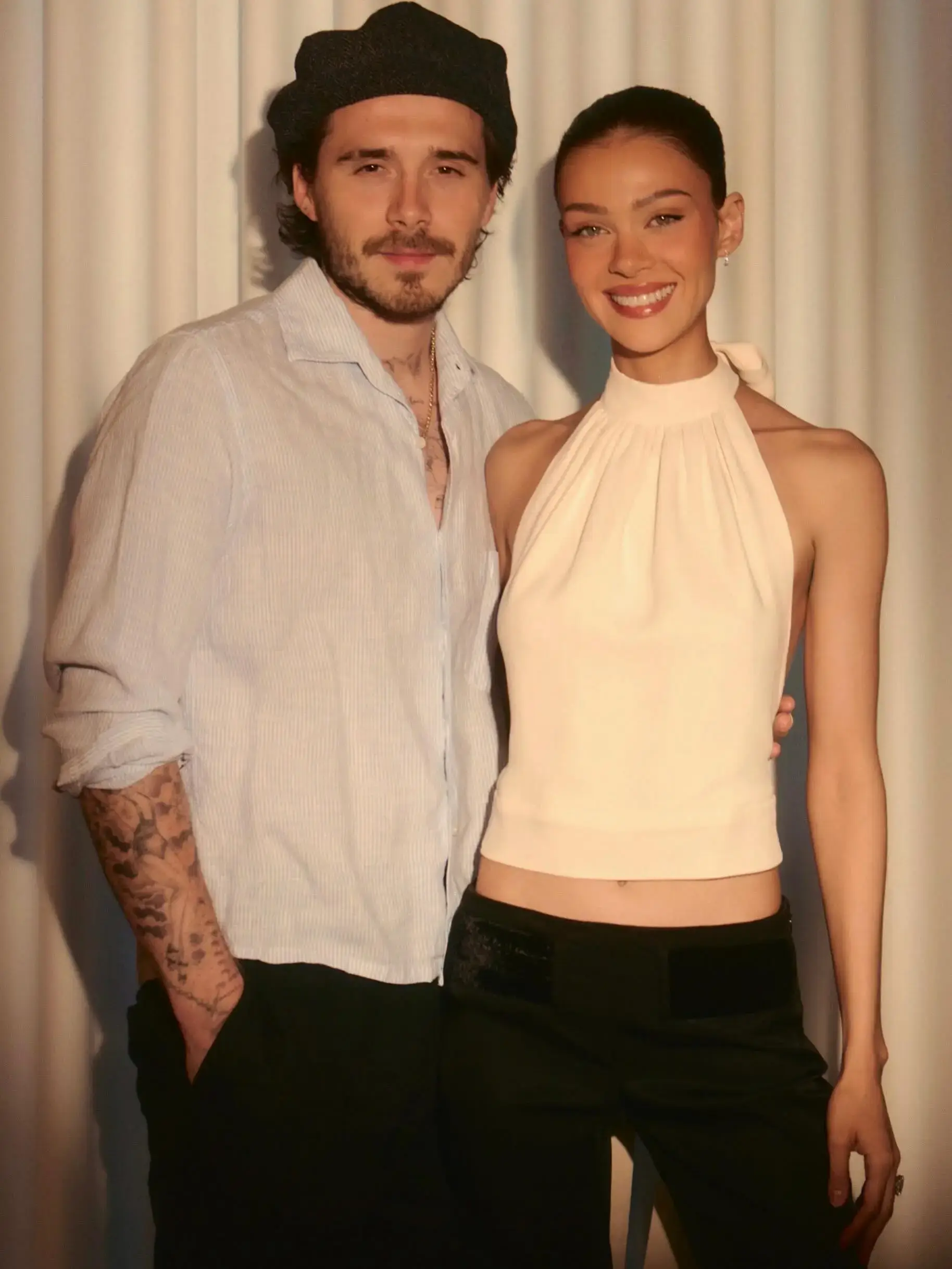 Brooklyn Beckham stands with his arm around his wife, Nicola Peltz, as they pose together against a white pleated curtain backdrop.