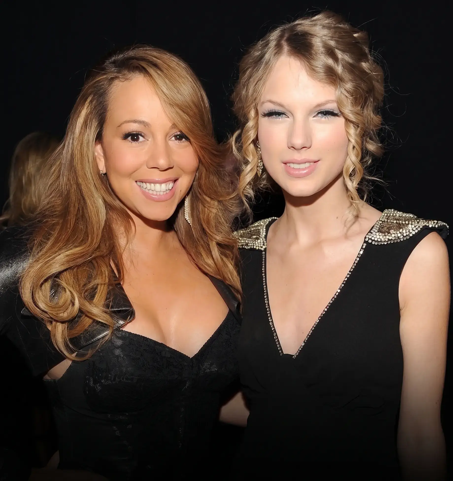 A close-up portrait of music icons Mariah Carey and Taylor Swift smiling together in black outfits against a dark background.