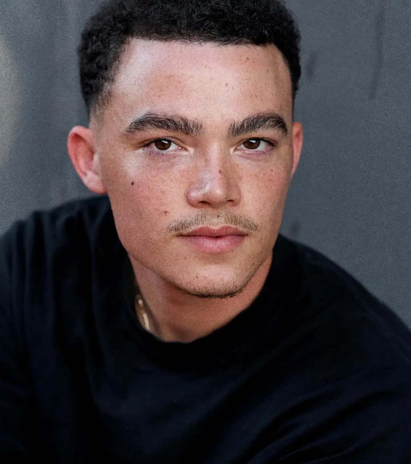 Actor Keanu Karim looking directly into the camera while wearing a black crew-neck shirt against a neutral grey background.