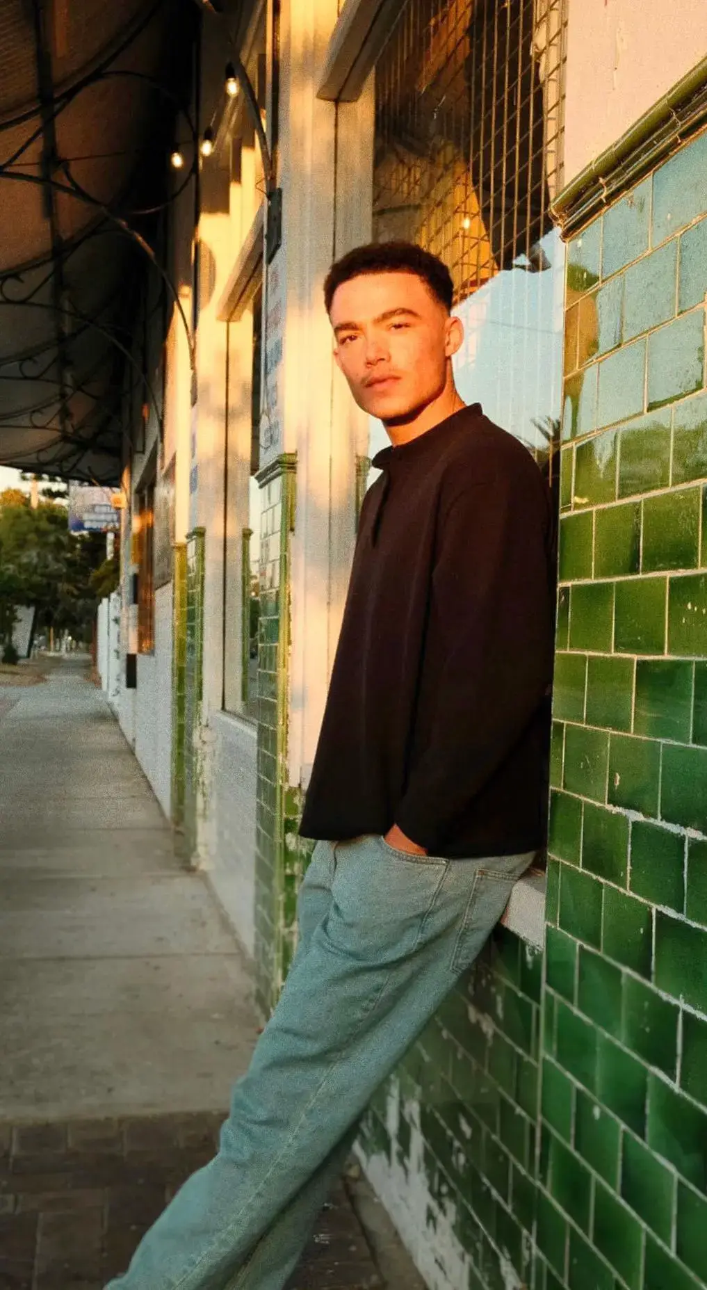 Actor Keanu Karim leans casually against a green-tiled wall outdoors while showing his full height in profile.
