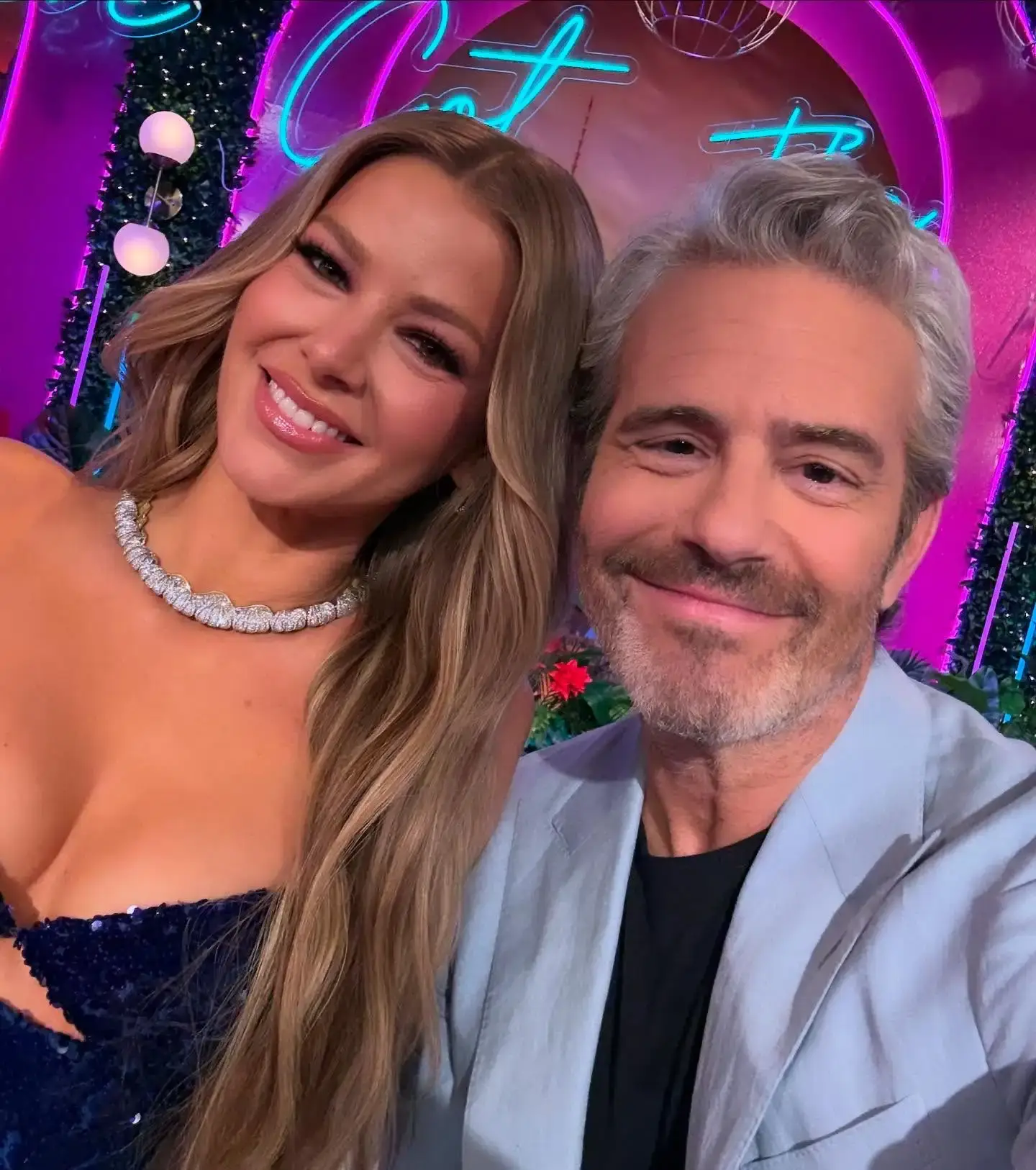 A close-up selfie of Ariana Madix in a sparkling dark blue dress and Andy Cohen in a light blue suit jacket smiling together on a brightly lit television set.
