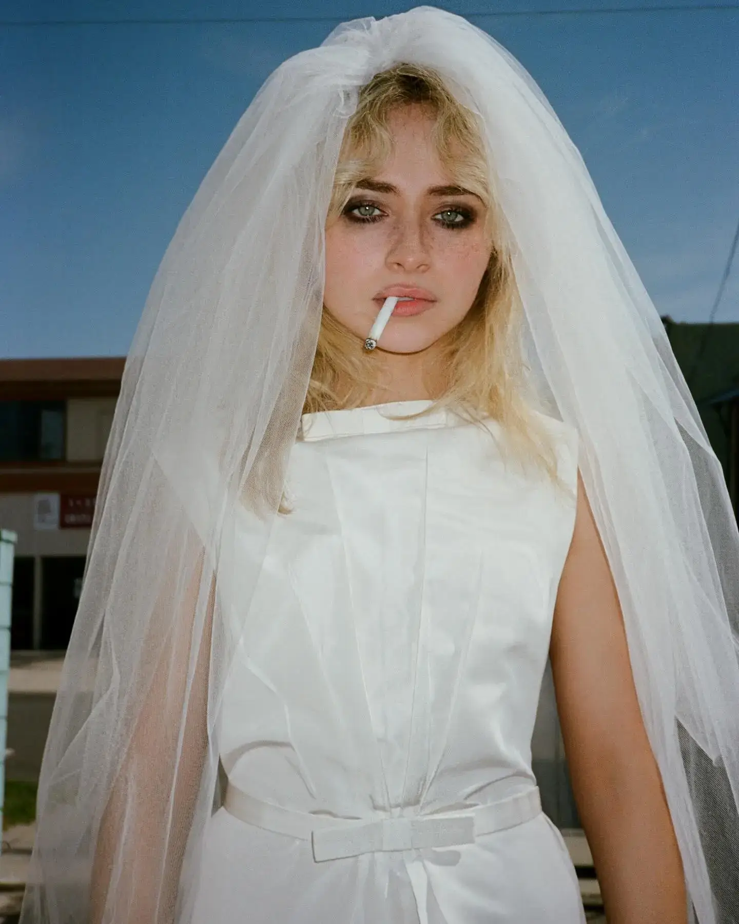 Singer Sabrina Carpenter poses in a white bridal gown and veil with a cigarette in her mouth against a blue sky.