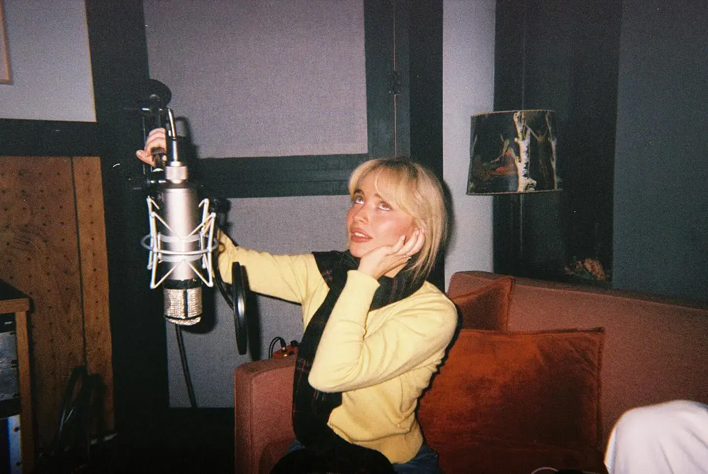 Singer Sabrina Carpenter looks up playfully while adjusting a studio microphone in a cozy recording booth.