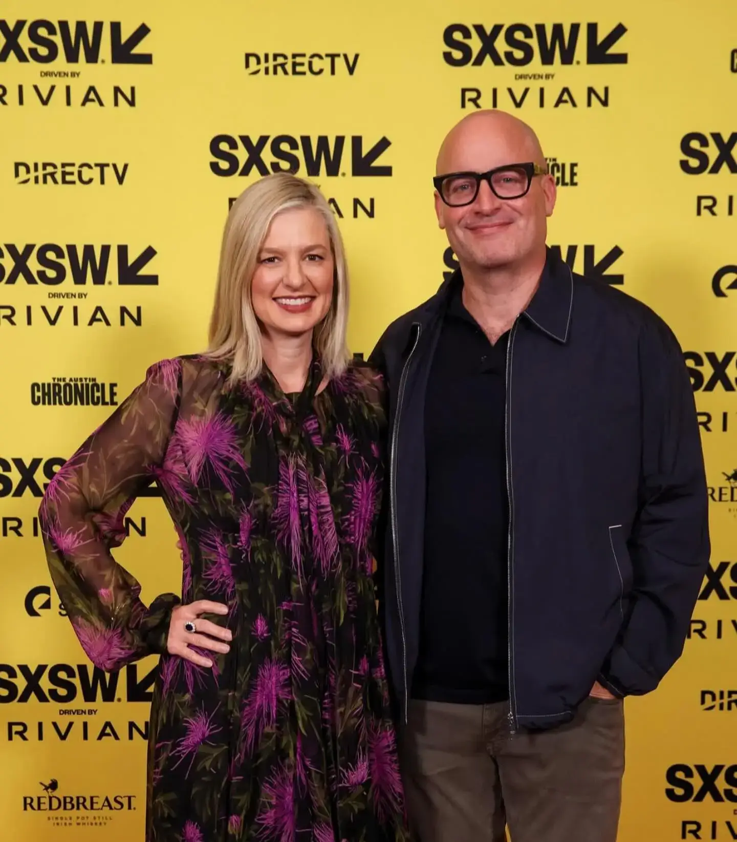 Christina Pazsitzky in a patterned black and purple dress and Tom Segura in a dark jacket stand smiling against a yellow SXSW step-and-repeat banner.