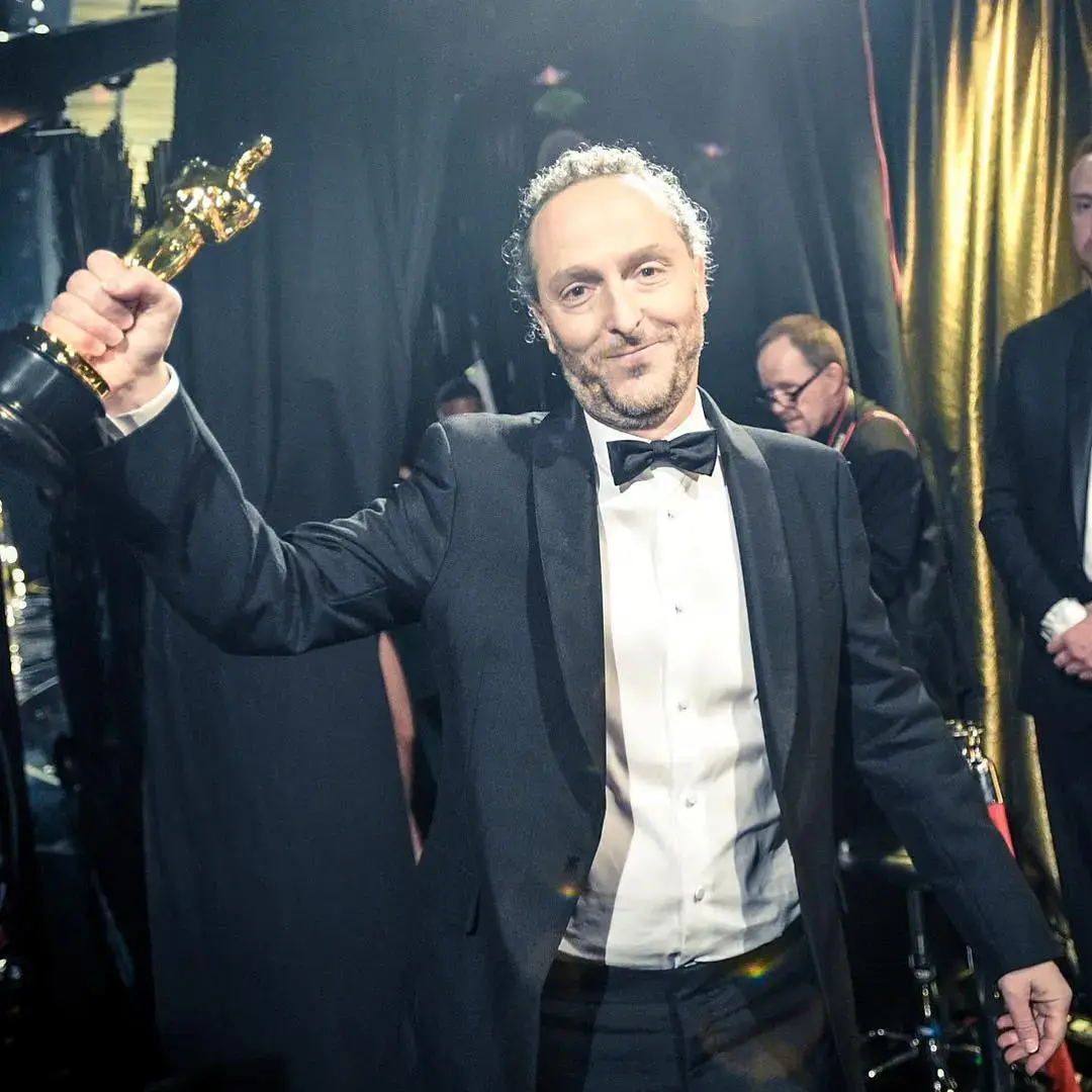 Cinematographer Emmanuel Lubezki holds up an Oscar trophy while smiling backstage at the Academy Awards in a black tuxedo and bow tie.