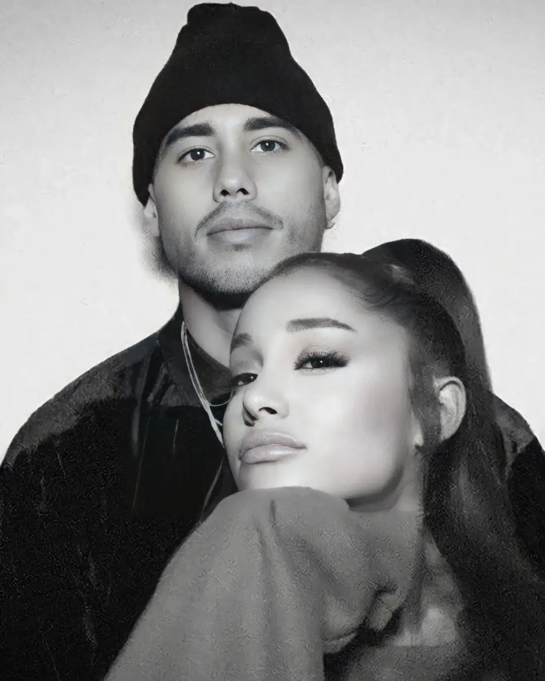 A desaturated close-up portrait of Ariana Grande resting her chin on her hand while Ricky Alvarez stands closely behind her wearing a black beanie.