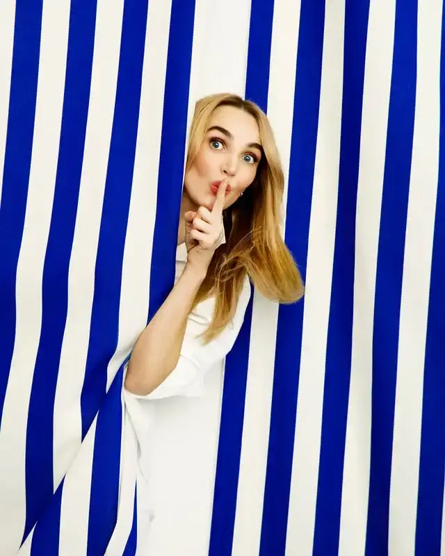 Actress Chloe Fineman peeks out from between bold blue and white vertically striped curtains while holding a finger to her lips.