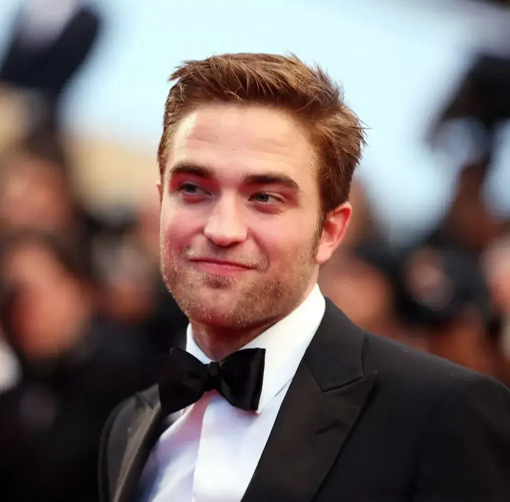 A close-up portrait of actor Robert Pattinson smiling slightly on a red carpet while wearing a black tuxedo and bow tie.