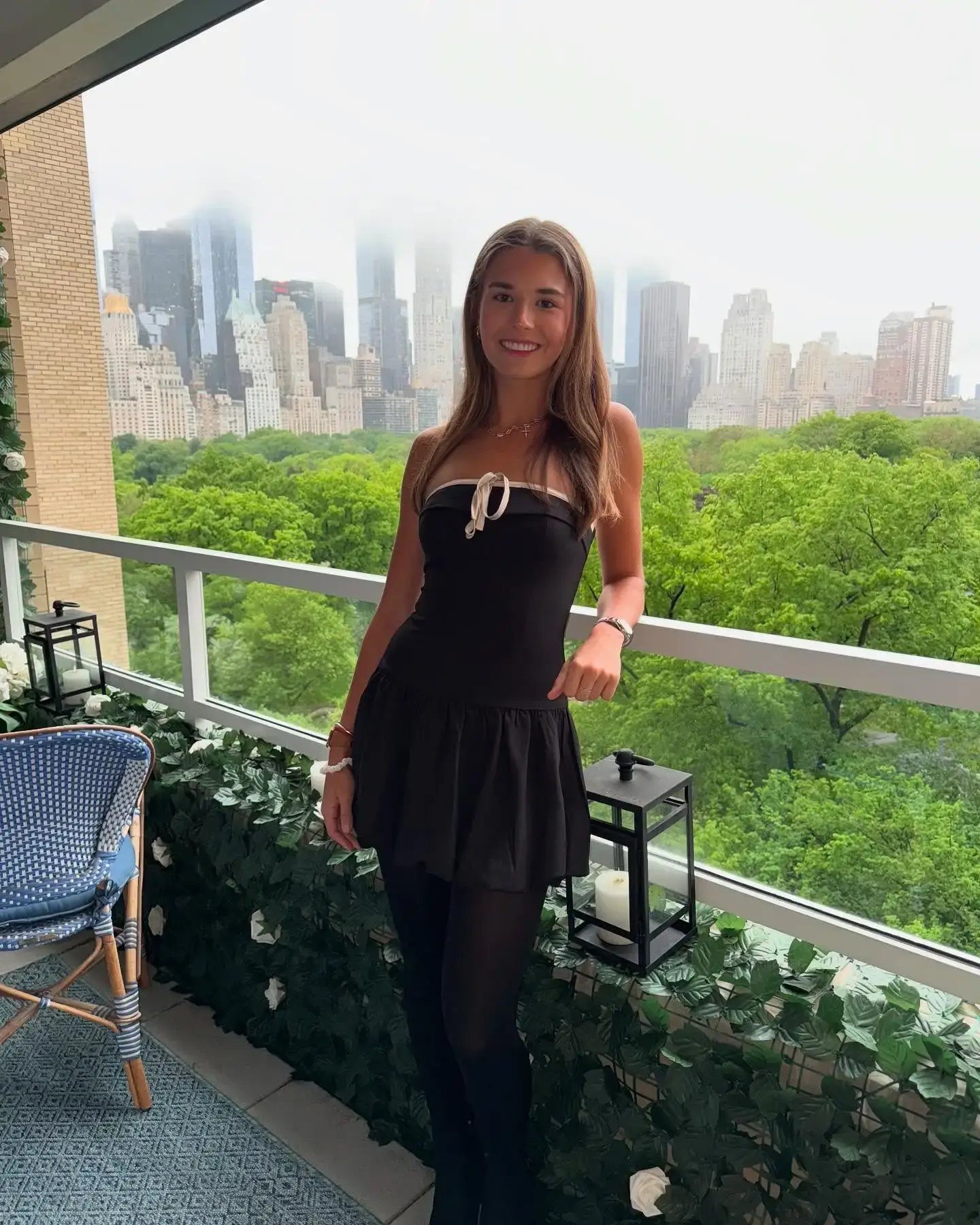 Kai Trump stands smiling on a balcony overlooking Central Park and the New York City skyline while wearing a black strapless mini dress and tights.
